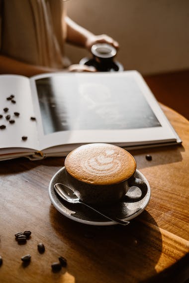 Sobre o canto arredondado de uma mesa de madeira, em primeiro plano uma xícara preta com café, cuja espuma desenha um coração. Adianta, um livro de gravuras aberto, com grãos de café sobre ele e sobre a mesa. Ao fundo, os braços de outra pessoa segurando uma segunda xícara com café.